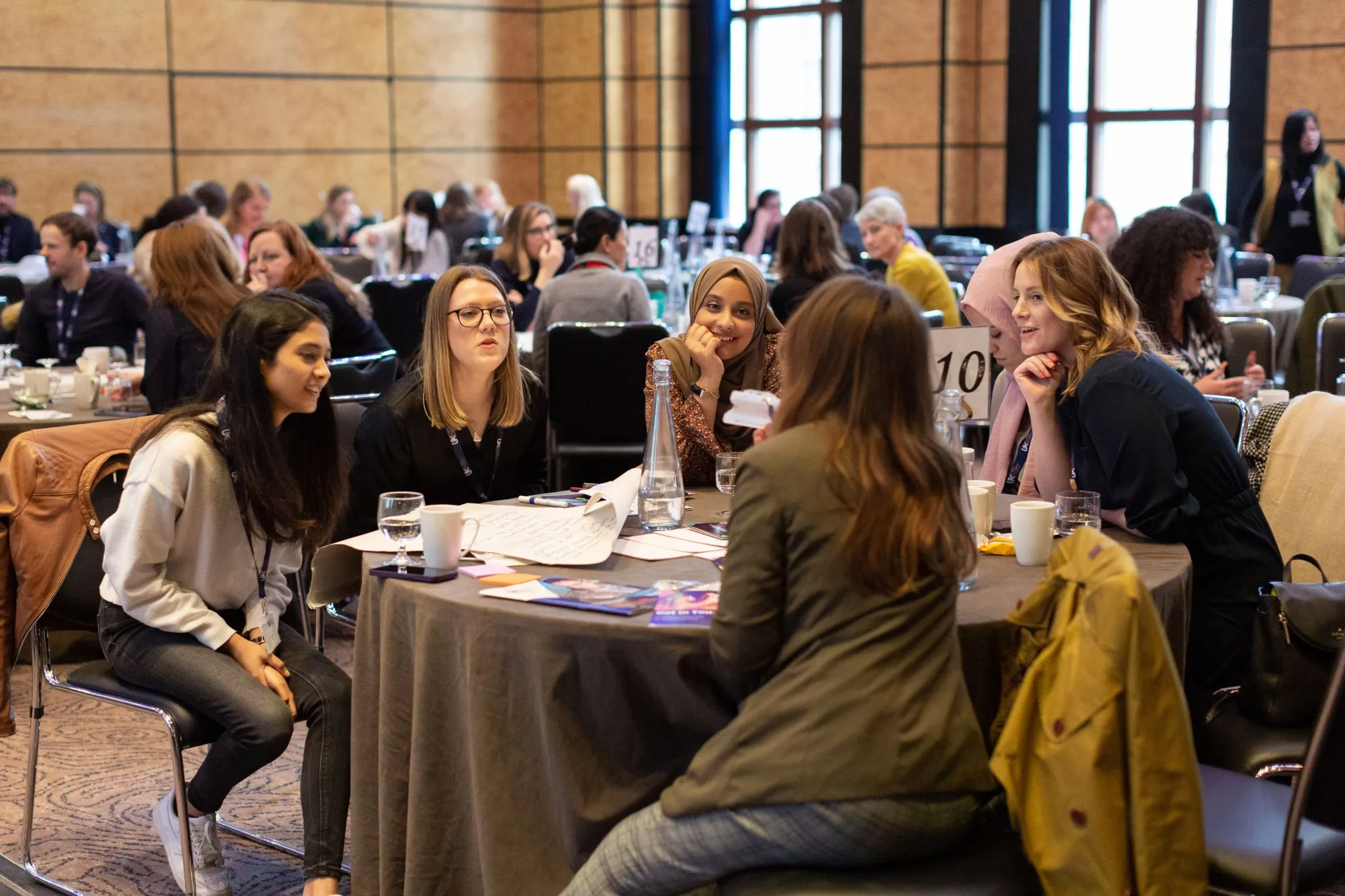 Lively roundtable discussion at a conference, with a diverse group of participants engaged in conversation around a table covered in notes, drinks, and booklets; other tables and attendees fill the background.