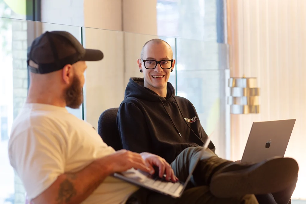Two people sitting in a relaxed workspace, chatting while using laptops; one wears a cap and white T-shirt, the other wears glasses and a black hoodie, smiling during the conversation.