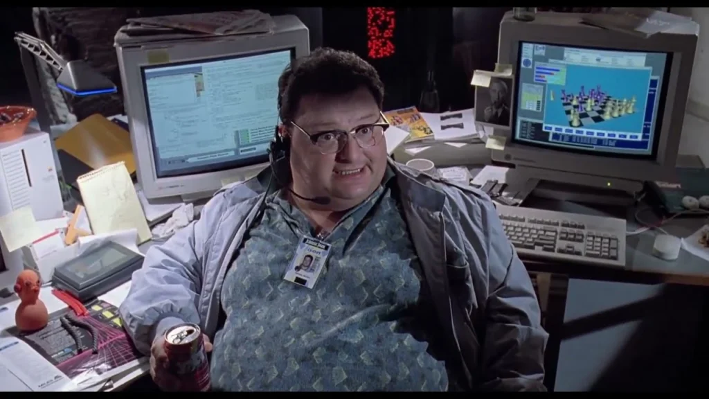 A cluttered 90s office scene with a person in glasses and a headset, wearing a Jurassic Park ID badge, sitting between two bulky CRT monitors—one displaying code and the other a 3D chess game—while holding a can of soda.