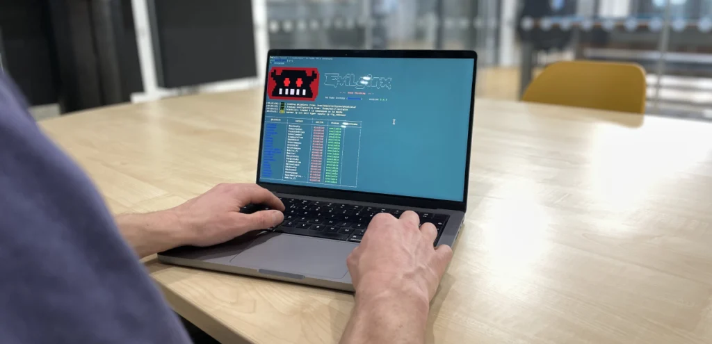 Close-up of hands typing on a laptop displaying a terminal interface for "Evilginx," a penetration testing tool, with a cartoon red skull icon and columns of status indicators; the scene takes place in a modern office meeting space.