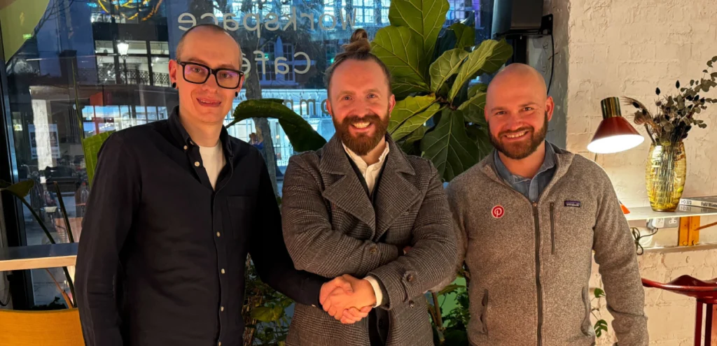 Three people standing indoors and smiling, with the two on the left shaking hands. They are positioned in front of large plants and a café window, suggesting a casual but celebratory setting.