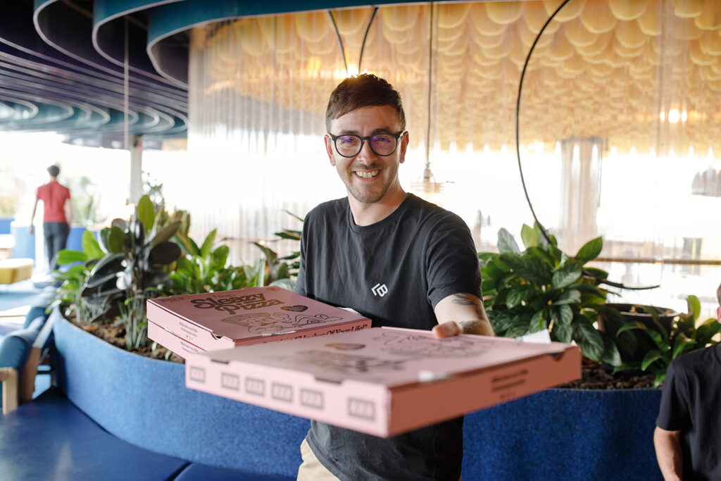 Smiling person in a black T-shirt offering two pink pizza boxes toward the camera, standing in a modern, plant-filled indoor space with curved blue planters and a warm yellow ceiling installation.