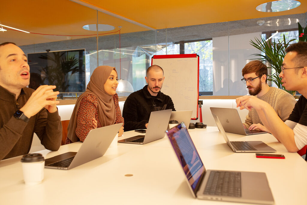 Six people from the agile Information Security Strategy team sit around a table in a modern meeting room, discussing work with laptops open and a whiteboard in the background.