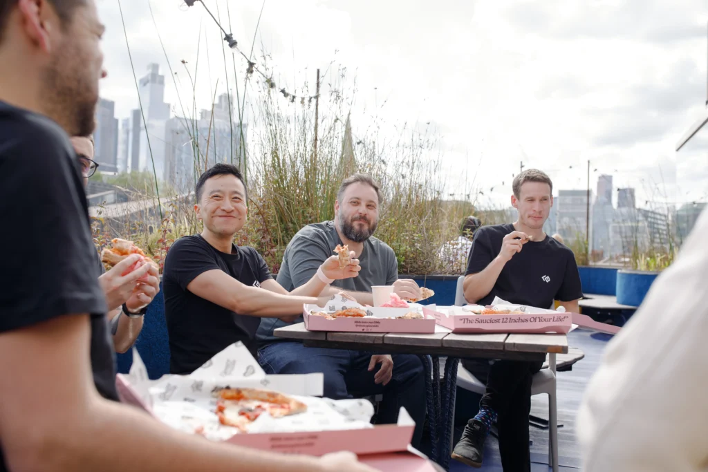 Group of people enjoying pizza outdoors on a rooftop terrace, with city buildings in the background; open pink pizza boxes on the table read “The Sauciest 12 Inches Of Your Life!” and the mood is cheerful and relaxed.