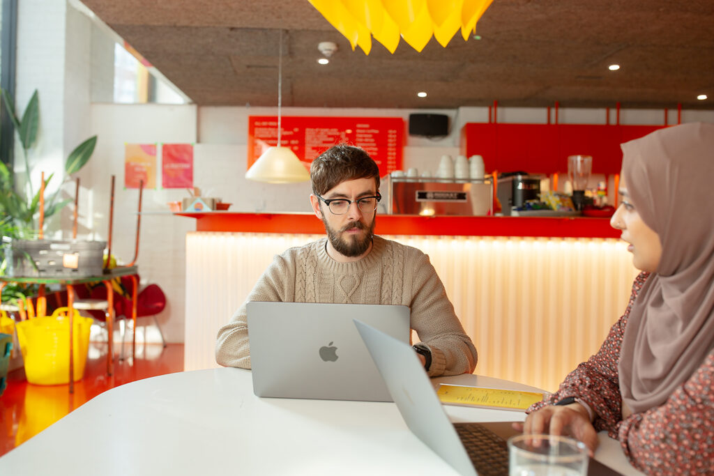 Two people sitting at a white table with laptops in a colourful café-style workspace, engaged in conversation. A bright red counter, warm lighting, and plants create a vibrant, relaxed atmosphere.