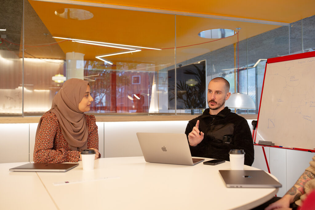 Two people sit at a round table in a modern office, engaged in discussion. One gestures while speaking, and the other listens attentively. Open laptops, takeaway coffee cups, and a whiteboard with diagrams are visible. Bright lighting and glass walls create a contemporary setting.