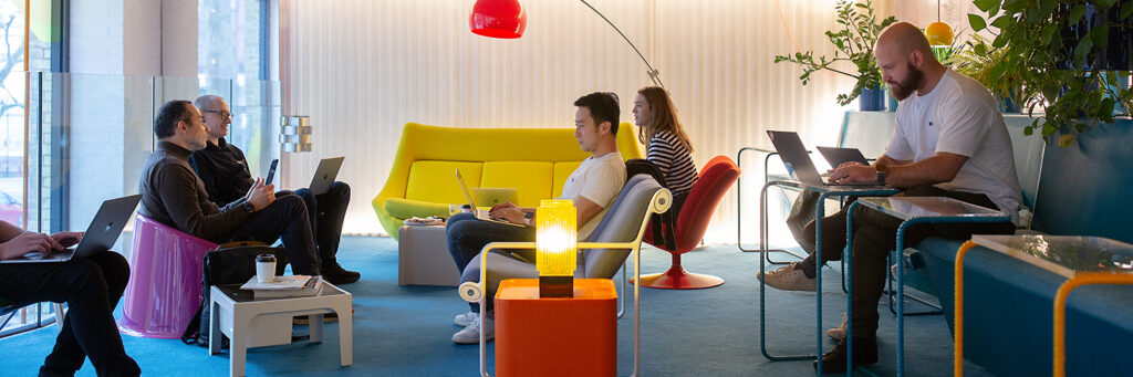 Group of people working on laptops in a colourful, modern lounge area with eclectic furniture, indoor plants, and a red overhead lamp.