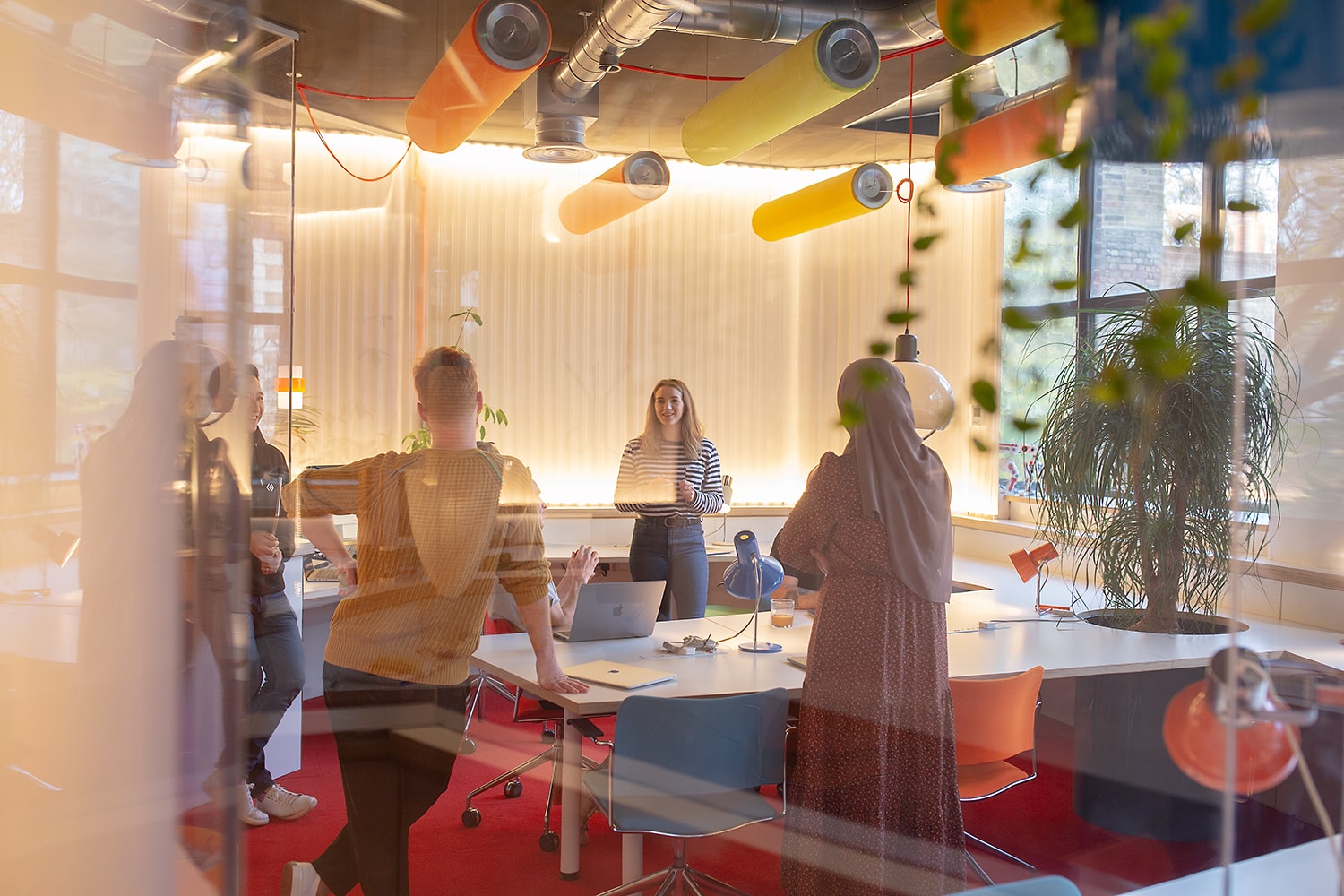 A small team gathers for a stand-up meeting in a bright, modern office with colourful acoustic ceiling panels, collaborative desks, and visible tech equipment—capturing a culture of agile teamwork and strategic IT planning.