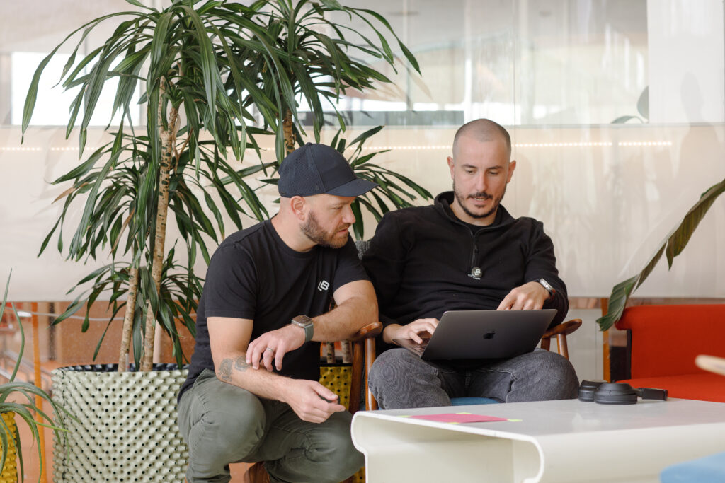 Two people sitting closely together in a relaxed office setting, discussing something on a laptop screen. One is seated while the other kneels beside, with large plants and a white table with headphones and notes in the foreground.