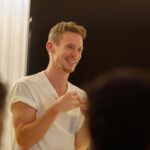 Person smiling while speaking in a warmly lit room, facing an audience with blurred heads in the foreground.