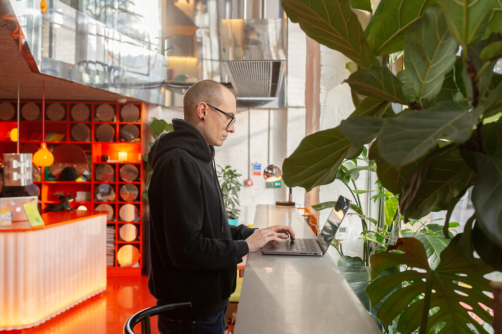 Person standing at a high counter by large windows, working on a laptop in a stylish, plant-filled space with vibrant red flooring and warm lighting from a nearby reception area.