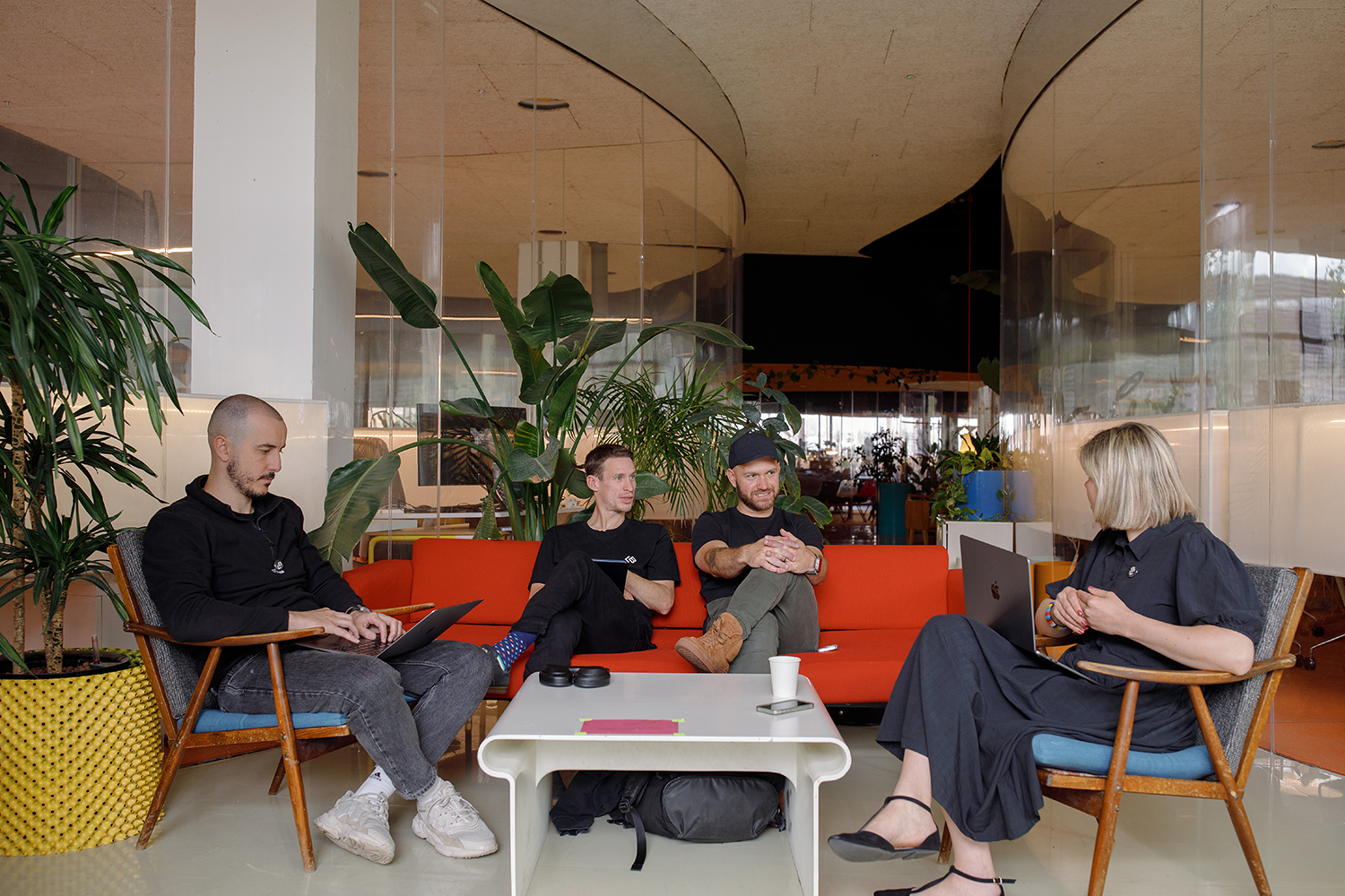 Four people sitting in a relaxed, plant-filled office lounge, engaged in discussion about Cyber Essentials certification with their laptops open and coffee cups on the table.