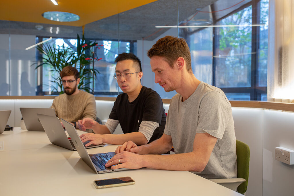 Three people seated at a white table working on laptops in a bright, modern office with large windows and indoor plants.