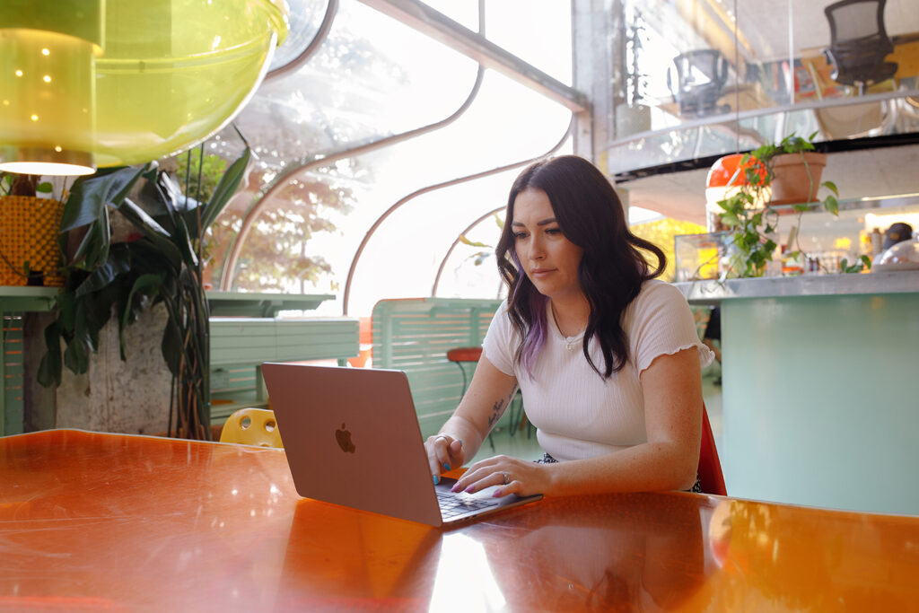 Person with long dark hair working on ISO 27001 certification a laptop at a bright orange table in a colourful, modern workspace with curved windows and plenty of plants.