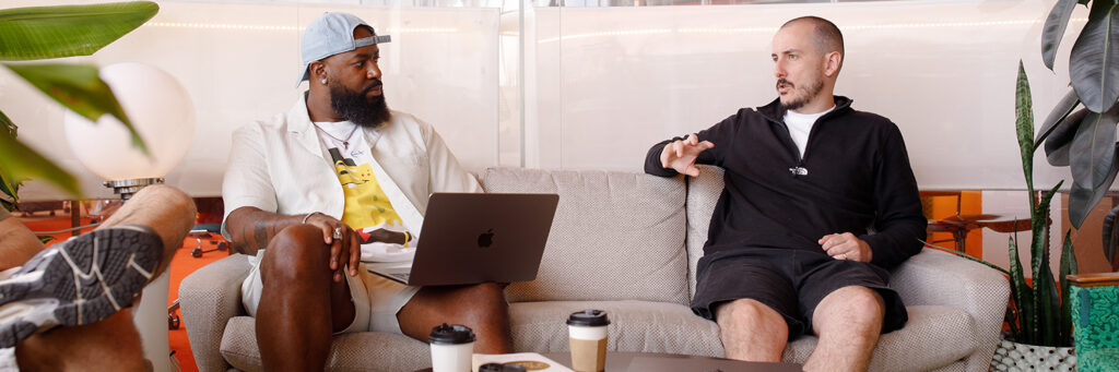 Two people sitting on a sofa in a casual, modern workspace, having a conversation with laptops and takeaway coffee cups on a round table in front of them.
