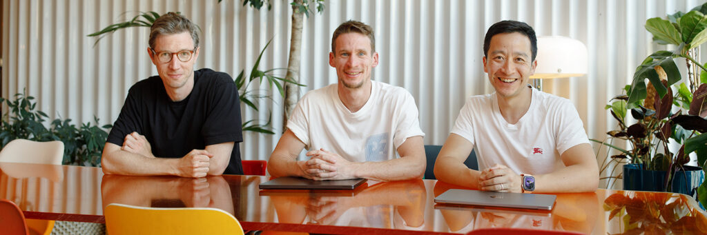 Three people seated at a colourful table in a bright, plant-filled room, smiling at the camera with laptops in front of them.