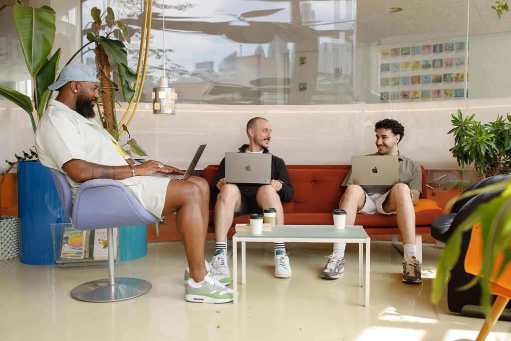 Two people sitting on a sofa in a casual, modern workspace, having a conversation about IT and security services with laptops and takeaway coffee cups on a round table in front of them. They are also talking about on and off boarding services here at LeftBrain.