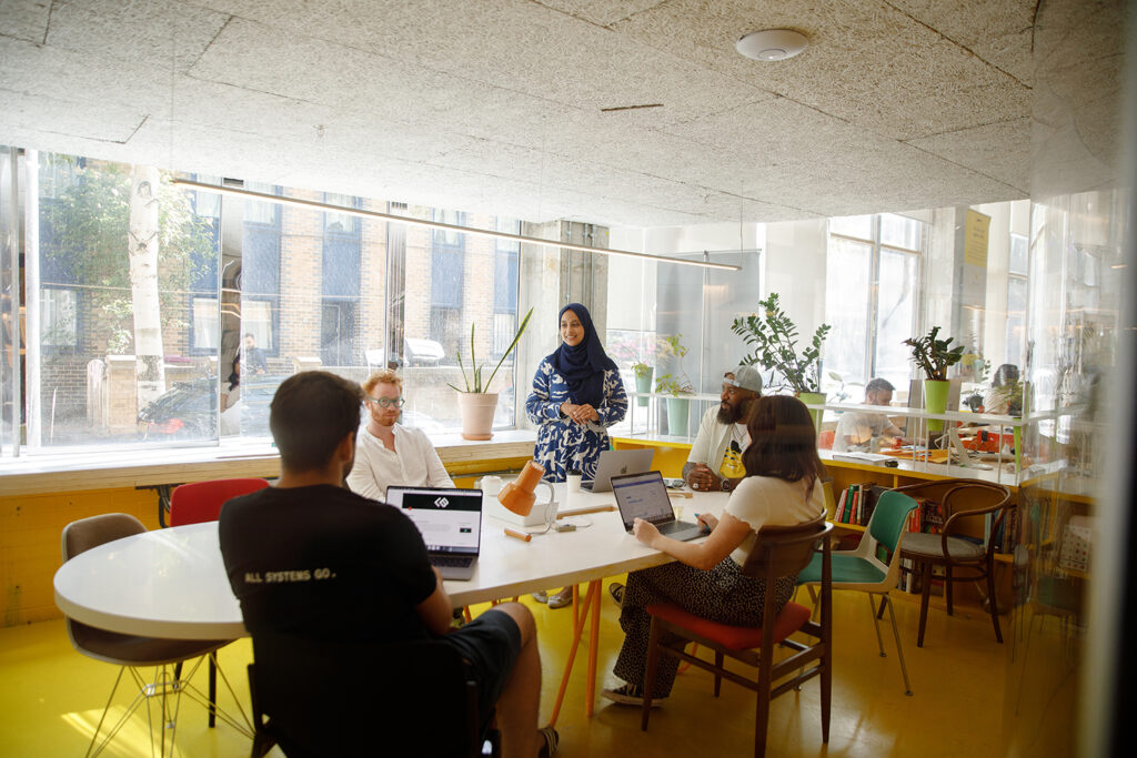 A group of five people in a brightly lit meeting room, with one person standing and speaking while the others sit around a table with laptops. The space has large windows, indoor plants, and a casual, modern design.