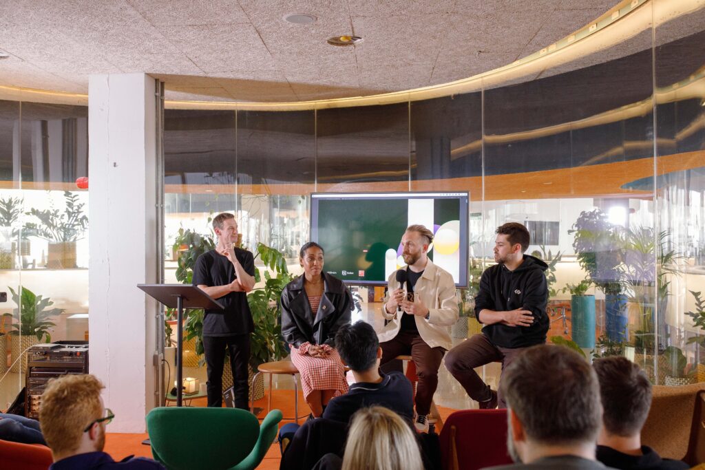 Alt text: Four speakers sit or stand in front of a screen during a panel discussion in a modern, plant-filled room, speaking to an audience seated in colourful chairs.