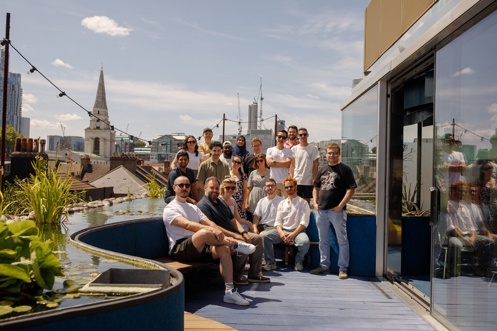 A group of people gathered on a rooftop terrace on a sunny day, posing for a photo beside a narrow pond with city buildings and a church spire visible in the background. They are celebrating being approved as a Security Reviewer to deliver HM Government's Secure Innovation Security reviews