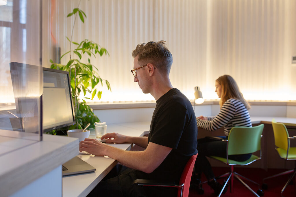 Two people working quietly at separate desks in a softly lit office, one focused on a desktop monitor and the other on a laptop, with plants and desk lamps nearby.