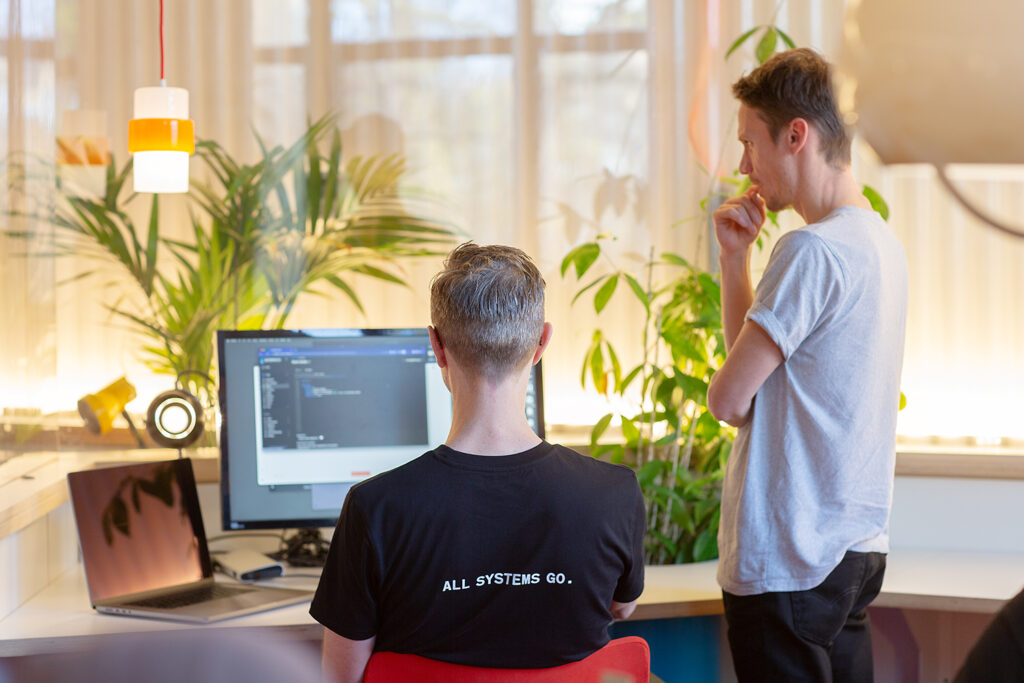 One person seated at a computer with visible code on the screen and “ALL SYSTEMS GO.” printed on their shirt, while another stands nearby thinking, surrounded by plants and warm lighting.
