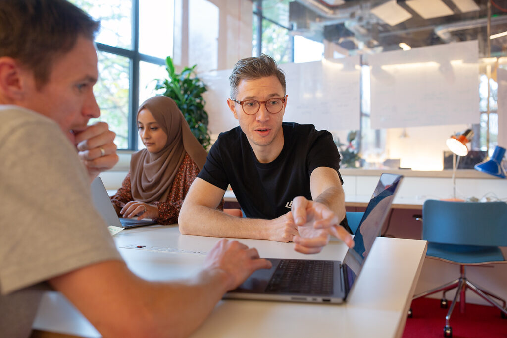 Three people seated around a white table in a modern office space, collaborating on laptops. One person gestures toward a screen while speaking, with whiteboards and greenery in the background.