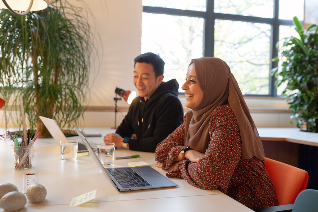 Two people seated at a desk in a bright office, smiling and engaged in conversation, with laptops, water glasses, and plants in the background.