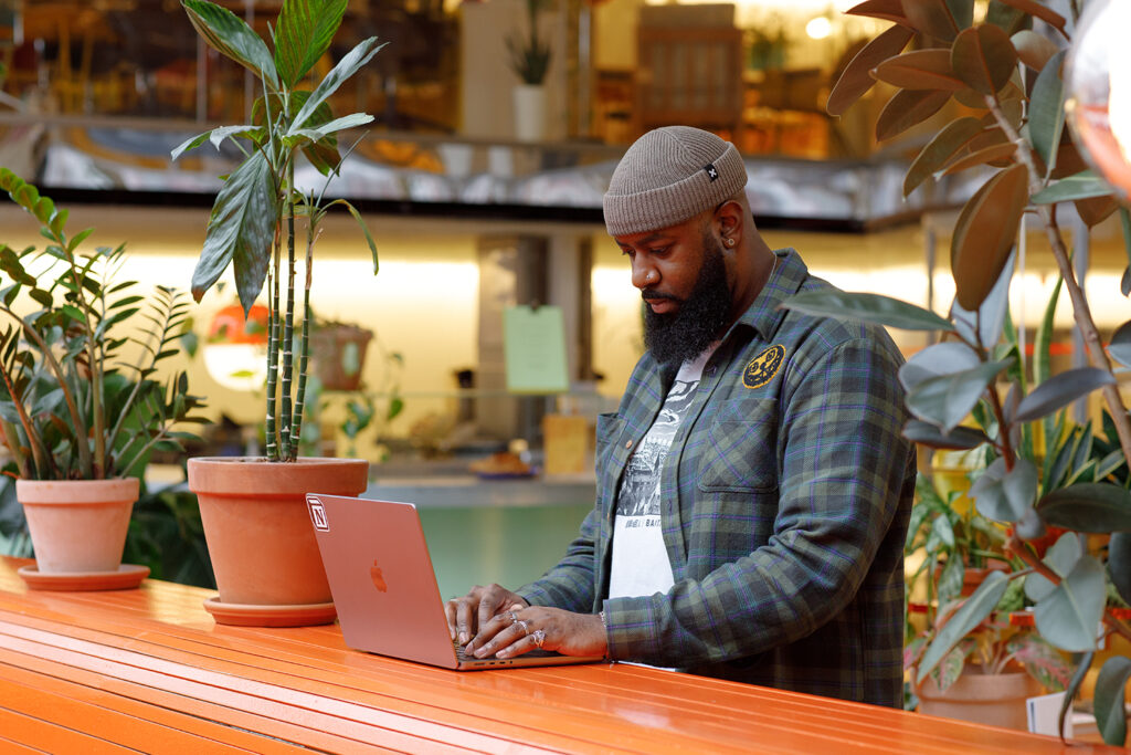 Alt text:
Person standing and typing on a laptop at a bright orange counter in a plant-filled workspace. The background features warm lighting, potted plants, and a modern interior with a relaxed atmosphere.