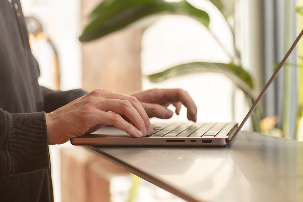 Close-up of hands typing on a laptop at a standing desk near a window, with soft natural light and green leaves in the background.
