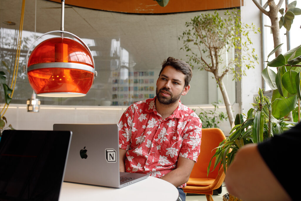 Person in a red floral shirt sitting at a table with a laptop, under a retro orange hanging lamp, in a modern indoor space with plants. They are discussing the benefits of ISO 27001 certification.