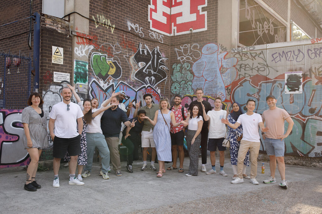 Group of LeftBrain staff posing cheerfully in front of a brick wall covered in colourful graffiti.
