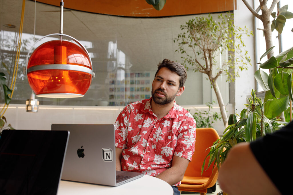 Person in a bright, plant-filled workspace, seated at a table with a laptop, listening during a conversation beneath a large red pendant light.
