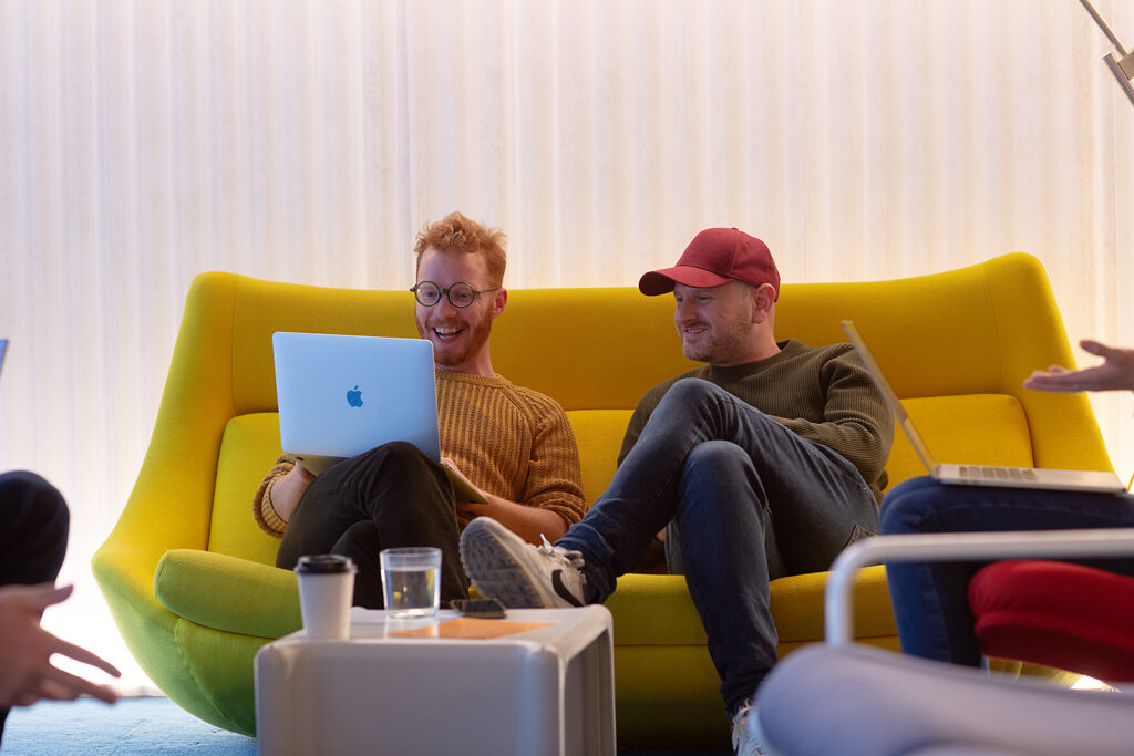 Two people sit on a bright yellow sofa, smiling at a laptop during an informal meeting, with coffee cups and notebooks nearby.
