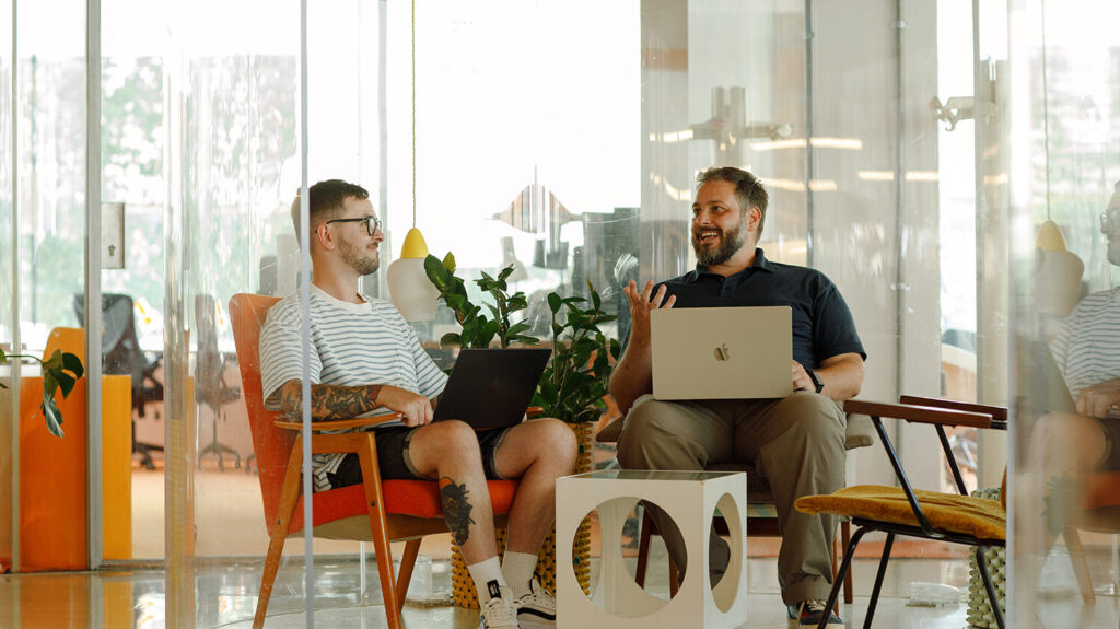 Two people sitting in a modern office lounge area, chatting and smiling while working on laptops, with plants and colourful furniture around them.