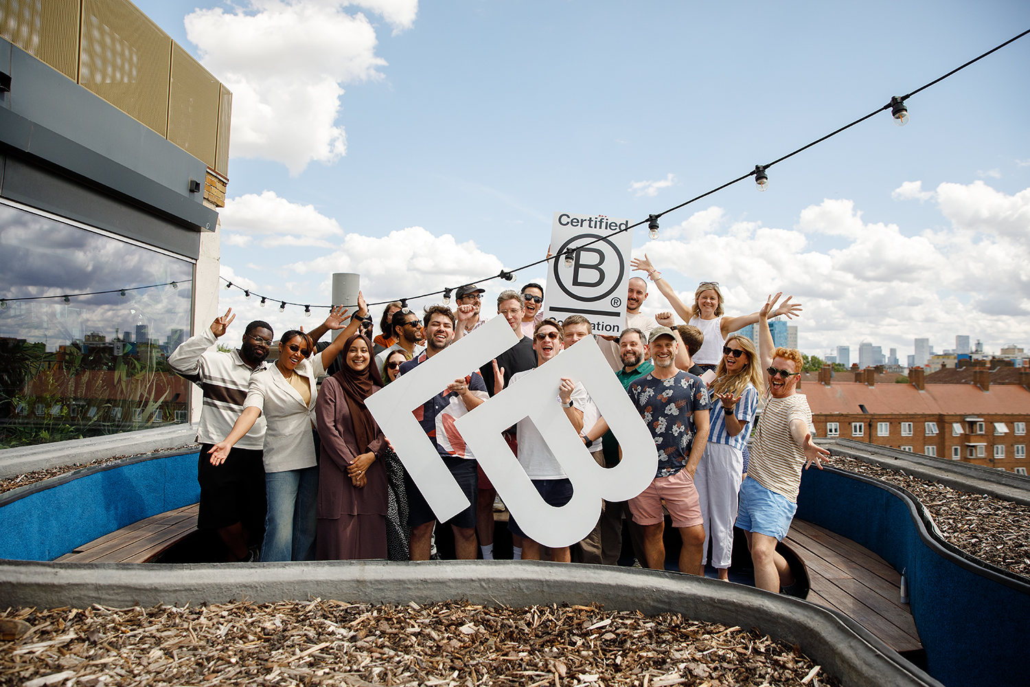 Team of colleagues gathered on the roof terrace in celebration holding a giant letter L and B and a sign with the B Corp logo on it.